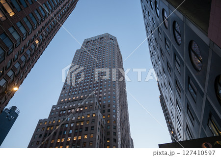 A towering skyscraper is framed by other high-rise buildings in New York City during early evening. The sky is clear as city lights begin to glow from office windows. A towering skyscraper is framed by other high-rise buildings in New York City during early evening. The sky is clear as city lights begin to glow from office windows. 127410597