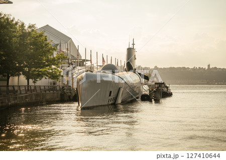 A black military submarine is moored at a pier lined with American flags, lit by warm evening sunlight. The calm river reflects the golden glow as trees and buildings fade into the hazy distance. 127410644