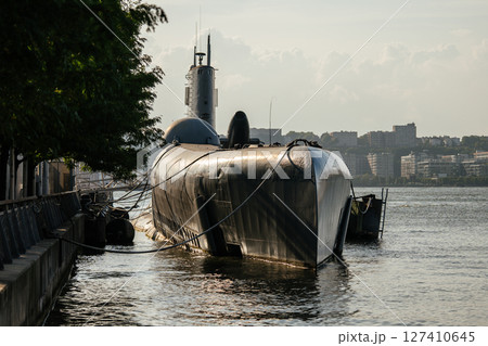 A black military submarine is docked at a quiet waterfront with mooring ropes securing it to the pier. The sun casts long reflections across the vessel's surface, with buildings visible in the 127410645