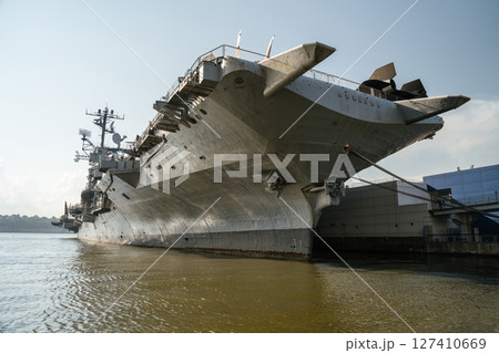 Side view of the USS carrier docked at a pier, showcasing its massive hull, anchor, and upper deck structures. The historic naval ship is now part of a museum in New York City. Side view of the USS carrier docked at a pier, showcasing its massive hull, anchor, and upper deck structures. The historic naval ship is now part of a museum in New York City. 127410669