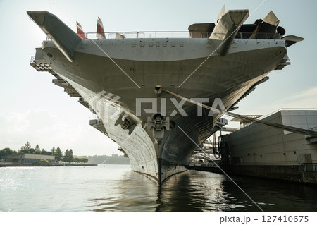 Frontal view of the USS aircraft carrier docked in New York, with the ship's bow reflecting on the calm water surface. The massive anchor and hull details are clearly visible. 127410675