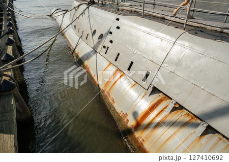 Detailed view of a submarine's hull showing streaks of rust and waterline marks. The vessel is docked at a wooden pier with ropes securing it above the water. 127410692