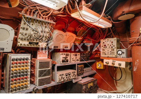 An array of analog controls, wires, and switches fills a submarine communication room. A warning sign, colored buttons, and monitoring devices are mounted across red metal walls. An array of analog controls, wires, and switches fills a submarine communication room. A warning sign, colored buttons, and monitoring devices are mounted across red metal walls. 127410717