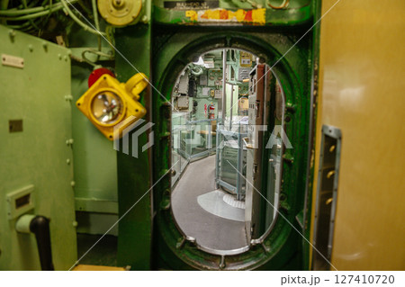 A green steel hatch opens into a submarine corridor with curved walls and glass barriers. A mounted yellow light is visible beside the entrance in the control section. A green steel hatch opens into a submarine corridor with curved walls and glass barriers. A mounted yellow light is visible beside the entrance in the control section. 127410720