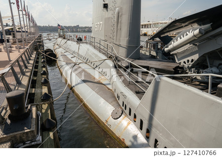 A preserved U.S. Navy submarine is docked at a waterfront museum with visible deck details and mooring ropes. American flags line the pier, and the city skyline can be seen in the background. 127410766
