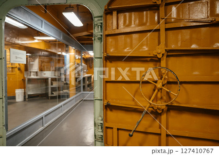 A vibrant orange watertight door with a large control wheel opens into a galley area behind glass. The image depicts part of a ship's internal structure, blending industrial design with utilitarian 127410767