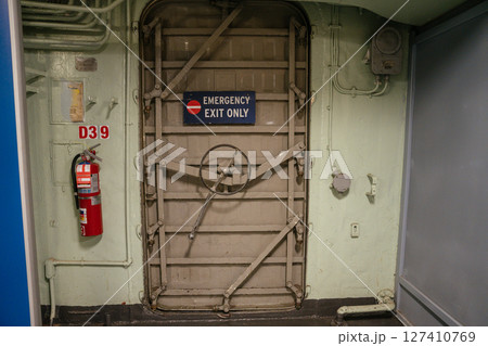 A secure emergency exit door with a wheel-lock mechanism is mounted on a ship or industrial wall. A red fire extinguisher is attached beside the hatch labeled. A secure emergency exit door with a wheel-lock mechanism is mounted on a ship or industrial wall. A red fire extinguisher is attached beside the hatch labeled. 127410769
