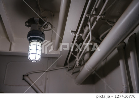 A glowing industrial-style ceiling lamp is mounted among painted metal pipes and conduit lines. The scene has a utilitarian and minimal aesthetic. A glowing industrial-style ceiling lamp is mounted among painted metal pipes and conduit lines. The scene has a utilitarian and minimal aesthetic. 127410770