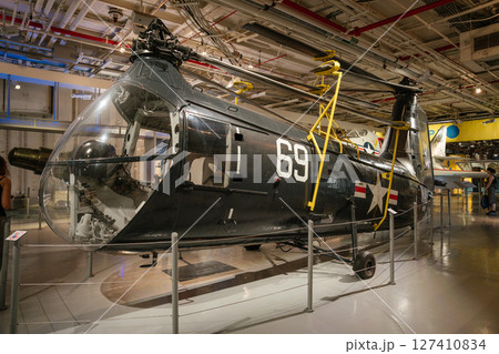 A vintage U.S. Navy helicopter with the number 69 is showcased inside an aviation museum. The aircraft is secured with barriers and surrounded by other historic aviation exhibits. 127410834