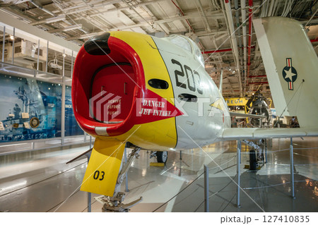 Close-up view of a jet intake labeled Danger Jet Intake on a U.S. Navy aircraft. The aircraft is exhibited inside an aviation museum with lighting and other planes visible. Close-up view of a jet intake labeled Danger Jet Intake on a U.S. Navy aircraft. The aircraft is exhibited inside an aviation museum with lighting and other planes visible. 127410835