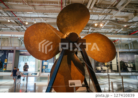 A large bronze ship propeller is mounted on a metal support inside a maritime museum. Museum visitors, including children, observe the impressive exhibit in a well-lit space. A large bronze ship propeller is mounted on a metal support inside a maritime museum. Museum visitors, including children, observe the impressive exhibit in a well-lit space. 127410839