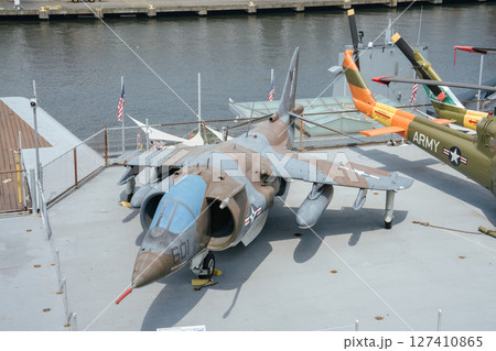A camouflaged military jet with a blue nose is displayed on the deck of a historic aircraft carrier. The jet is positioned near helicopters and fenced off from the river in the background. A camouflaged military jet with a blue nose is displayed on the deck of a historic aircraft carrier. The jet is positioned near helicopters and fenced off from the river in the background. 127410865