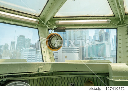 A vintage analog clock is mounted on a green steel beam inside a ship's control bridge. Through the wide windows, a modern city skyline with tall buildings is clearly visible. A vintage analog clock is mounted on a green steel beam inside a ship's control bridge. Through the wide windows, a modern city skyline with tall buildings is clearly visible. 127410872