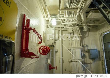 A close-up of a red valve and an empty extinguisher holder on a wall inside a ship's interior. Numerous cables are neatly bundled above, showing an industrial and functional design. A close-up of a red valve and an empty extinguisher holder on a wall inside a ship's interior. Numerous cables are neatly bundled above, showing an industrial and functional design. 127410890