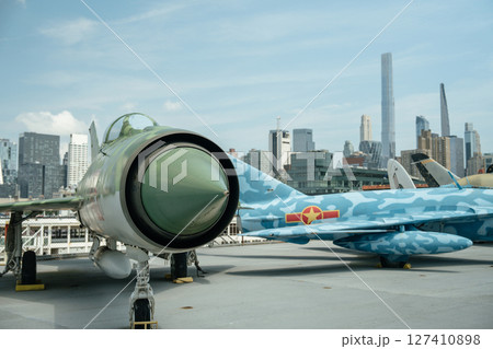 A green Soviet-era fighter jet is displayed on an outdoor deck with a blue camouflage aircraft beside it. The New York City skyline rises in the background, creating a contrast between military 127410898
