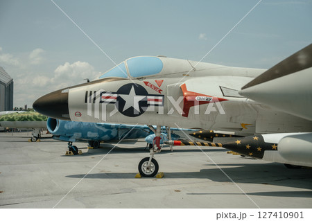 A detailed side view of a historic U.S. Navy fighter jet is shown parked on a carrier deck. The aircraft features missile mounts, rescue markings, and a military insignia with clear warning labels. 127410901