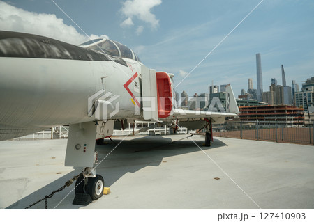 A close-up angle of a military jet fighter shows its cockpit, engine intake, and landing gear. The aircraft is parked on a rooftop with a city skyline in the background. 127410903