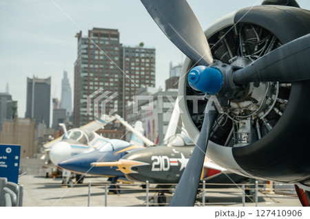 A close-up of a vintage military propeller engine is displayed with other aircraft and New York City skyscrapers in the background. The black propeller blades and exposed engine parts dominate the A close-up of a vintage military propeller engine is displayed with other aircraft and New York City skyscrapers in the background. The black propeller blades and exposed engine parts dominate the 127410906
