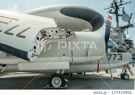 A detailed view of a white military propeller plane shows an open engine maintenance panel and exposed mechanical parts. The aircraft is parked on a tarmac near a naval radar tower. A detailed view of a white military propeller plane shows an open engine maintenance panel and exposed mechanical parts. The aircraft is parked on a tarmac near a naval radar tower. 127410908