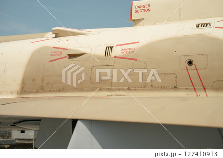 Close-up view of a beige military aircraft showing red warning labels and vent systems. Markings such as Danger Suction and Danger Hot Air are clearly visible on the fuselage. Close-up view of a beige military aircraft showing red warning labels and vent systems. Markings such as Danger Suction and Danger Hot Air are clearly visible on the fuselage. 127410913