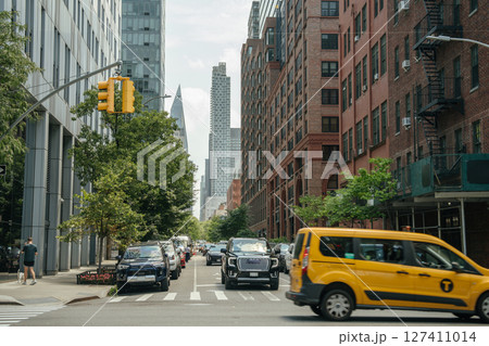 A yellow New York City taxi passes through a modern downtown street lined with parked cars and tall buildings. The background includes glass skyscrapers and red brick architecture, capturing a typical A yellow New York City taxi passes through a modern downtown street lined with parked cars and tall buildings. The background includes glass skyscrapers and red brick architecture, capturing a typical 127411014