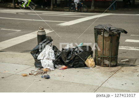 Black garbage bags and loose trash lie next to an overflowing street bin on a concrete sidewalk. A fire hydrant and urban crosswalk mark the busy city setting. Black garbage bags and loose trash lie next to an overflowing street bin on a concrete sidewalk. A fire hydrant and urban crosswalk mark the busy city setting. 127411017