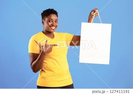 Great Sales. Happy Black Woman Showing Shopping Bag Pointing Finger At Blank White Shopper Handbag Standing Over Blue Studio Background, Smiling To Camera. Discounts Offer Advertisement. Mockup 127411191