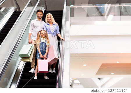 A happy family, a father, mother, and young daughter, are riding up an escalator in a modern shopping mall. They are all smiling and holding shopping bags, copy space A happy family, a father, mother, and young daughter, are riding up an escalator in a modern shopping mall. They are all smiling and holding shopping bags, copy space 127411194