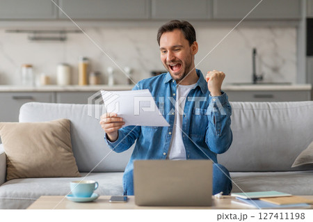 A man is sitting on a couch in a modern living room, holding a piece of paper and smiling widely. He has his right fist raised in the air in a gesture of celebration. He is likely reading good news 127411198