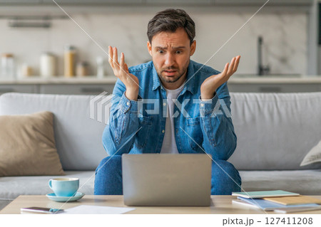 A man sits on a couch in his living room, looking confused as he stares at his laptop. His arms are raised in a gesture of frustration. A cup of coffee sits on a table in front of him. A man sits on a couch in his living room, looking confused as he stares at his laptop. His arms are raised in a gesture of frustration. A cup of coffee sits on a table in front of him. 127411208