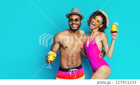 Black man and woman are smiling and enjoying summer cocktails in front of a blue background. The man is wearing sunglasses and a hat while the woman is wearing sunglasses and a swimsuit. Black man and woman are smiling and enjoying summer cocktails in front of a blue background. The man is wearing sunglasses and a hat while the woman is wearing sunglasses and a swimsuit. 127411233