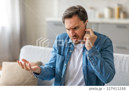A man sits on a couch in a living room, frowning and gesturing with his hand while talking on a phone. He is frustrated and angry, and it seems like he is in the middle of a heated conversation. A man sits on a couch in a living room, frowning and gesturing with his hand while talking on a phone. He is frustrated and angry, and it seems like he is in the middle of a heated conversation. 127411288