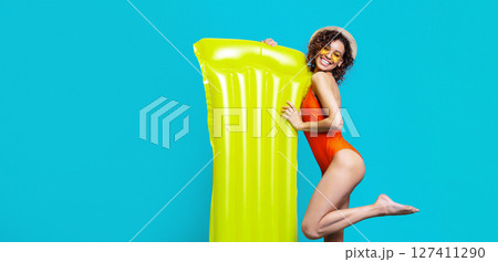 African American young woman in a red swimsuit, sunglasses, and a straw hat smiles while holding a large yellow inflatable raft against a blue background African American young woman in a red swimsuit, sunglasses, and a straw hat smiles while holding a large yellow inflatable raft against a blue background 127411290
