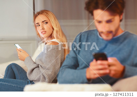 Infidelity. Wife Catching Cheating Husband Texting On Cellphone With Another Woman Sitting On Sofa At Home. Selective Focus 127411311