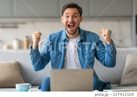 A man with a wide, joyful smile sits on a couch, his hands raised in victory. He is wearing a blue denim shirt and is seated in front of a laptop computer. A man with a wide, joyful smile sits on a couch, his hands raised in victory. He is wearing a blue denim shirt and is seated in front of a laptop computer. 127411329