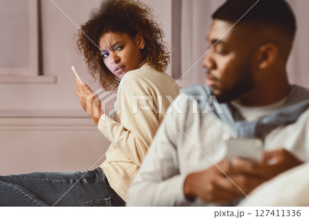 Afro woman suspecting husband in betrayal, using phone and looking at man 127411336