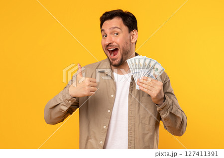 A man is holding a fan of cash in his right hand and giving a thumbs up with his left hand. He is smiling and looks excited while standing in front of a bright yellow background. A man is holding a fan of cash in his right hand and giving a thumbs up with his left hand. He is smiling and looks excited while standing in front of a bright yellow background. 127411391