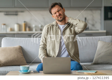 A man sits on a couch, working on a laptop. He is holding his neck and appears to be in pain. The couch is in a living room setting. 127411395