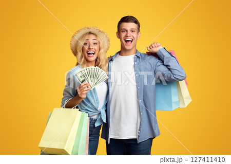 Tax Free Concept. Excited Young Couple Holding Paper Bags And Bunch Of Dollars Happy About Cashback After Successful Shopping Abroad, Panorama With Copy Space 127411410
