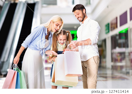 A young girl is standing between her parents, all three looking down at a pile of shopping bags. The girl is smiling, and the parents are smiling as they look at the contents of the bags. 127411414