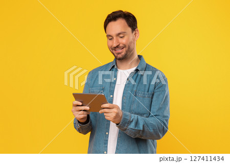 A man wearing a denim shirt stands in front of a bright yellow background, holding a tablet computer in his hands and looking down at the screen. He is smiling as he interacts with the tablet. 127411434
