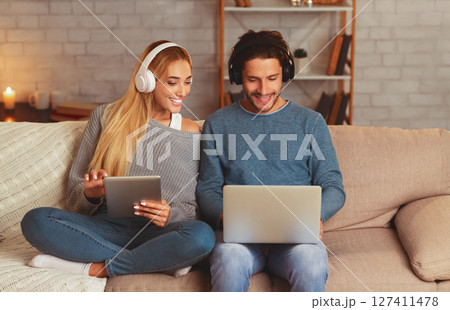 Modern Lifestyle. Smiling Couple Using Laptop And Tablet Computers Wearing Headphones Sitting On Couch At Home In Evening 127411478