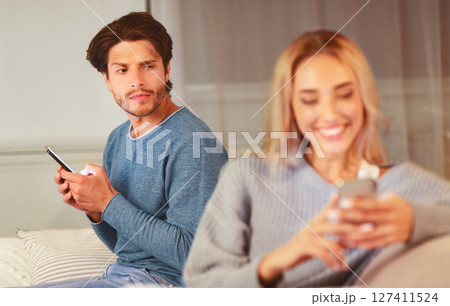 Infidelity. Suspicious Man Watching His Girlfriend Texting On Phone Suspecting Unfaithfulness Sitting On Sofa At Home. Selective Focus Infidelity. Suspicious Man Watching His Girlfriend Texting On Phone Suspecting Unfaithfulness Sitting On Sofa At Home. Selective Focus 127411524