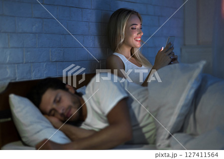 Cheating Wife Chatting On Cellphone While Her Husband Sleeping In Bedroom At Night. Low Light, Selective Focus Cheating Wife Chatting On Cellphone While Her Husband Sleeping In Bedroom At Night. Low Light, Selective Focus 127411564