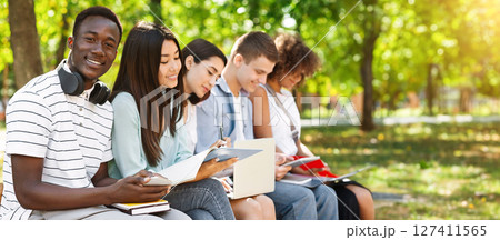 Smiling African American Student Guy Studying Outdoors With His University Friends, Holding Smartphone And Smiling At Camera, Selective Focus Smiling African American Student Guy Studying Outdoors With His University Friends, Holding Smartphone And Smiling At Camera, Selective Focus 127411565