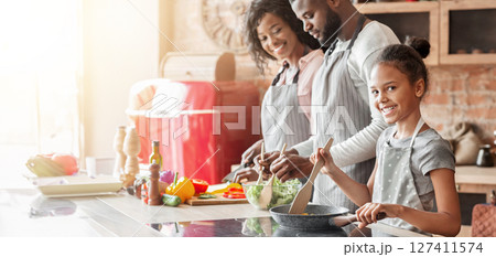 Smiling african little girl helping parents cooking healthy dinner in kitchen, frying vegetables Smiling african little girl helping parents cooking healthy dinner in kitchen, frying vegetables 127411574