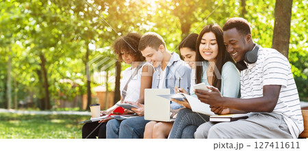 Education concept. Cheerful university students preparing for exam, talking and drinking coffee outdoors, sitting on bench in campus, free space Education concept. Cheerful university students preparing for exam, talking and drinking coffee outdoors, sitting on bench in campus, free space 127411611