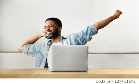 A man stretches back in his chair at his desk, smiling and looking relaxed. His laptop sits open in front of him. 127411675