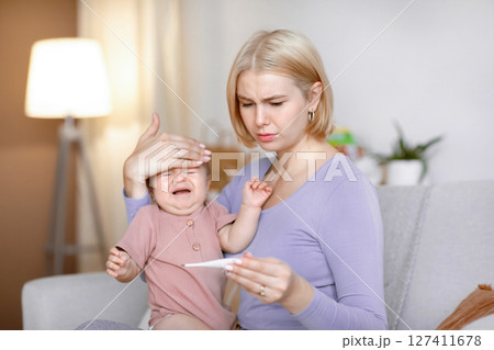 Worried woman young mother checking temperature to her sick crying baby, using electronic thermometer, home interior. Little kid got fever, troubled mom looking at thermometer. Infant health problems Worried woman young mother checking temperature to her sick crying baby, using electronic thermometer, home interior. Little kid got fever, troubled mom looking at thermometer. Infant health problems 127411678