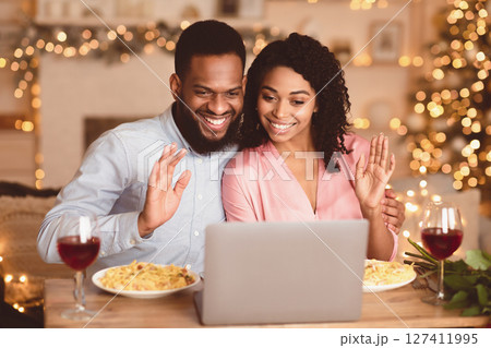 Virtual Party. Black couple sitting at the table, having dinner, drinking wine and having video call with parents or friends on laptop, waving to webcam. Stay home, quarantine celebration of event Virtual Party. Black couple sitting at the table, having dinner, drinking wine and having video call with parents or friends on laptop, waving to webcam. Stay home, quarantine celebration of event 127411995